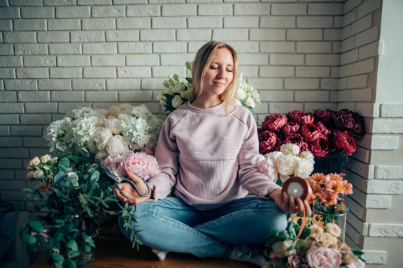 Woman meditating among colorful flower arrangements in a bright indoor setting.